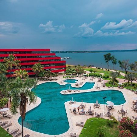 Royal Tulip Brasilia Alvorada Hotel Exterior photo The photo shows a scenic view of a resort or hotel with a striking red building in the background. In the foreground, there is a large, uniquely shaped swimming pool surrounded by palm trees and lounge chairs. The pool appears inviting, with clear wa