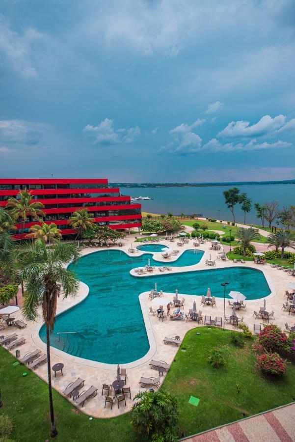 Royal Tulip Brasilia Alvorada Hotel Exterior photo The photo shows a scenic view of a resort or hotel with a striking red building in the background. In the foreground, there is a large, uniquely shaped swimming pool surrounded by palm trees and lounge chairs. The pool appears inviting, with clear wa