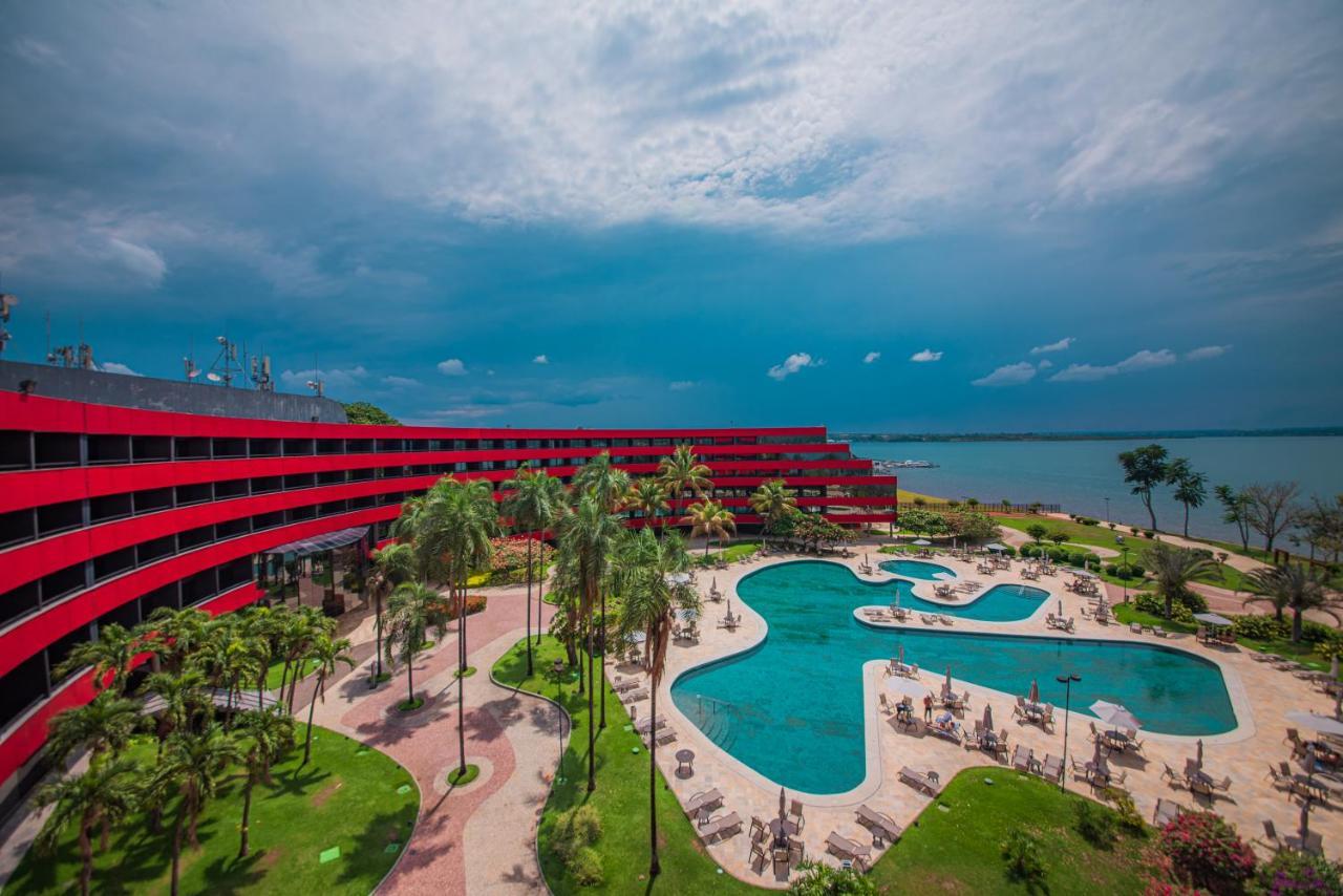 Royal Tulip Brasilia Alvorada Hotel Exterior photo The photo shows a modern hotel building with a striking red exterior, surrounded by lush greenery. In the foreground, there is a large, creatively shaped swimming pool, with lounge chairs arranged around it. The backdrop features a body of water, pos