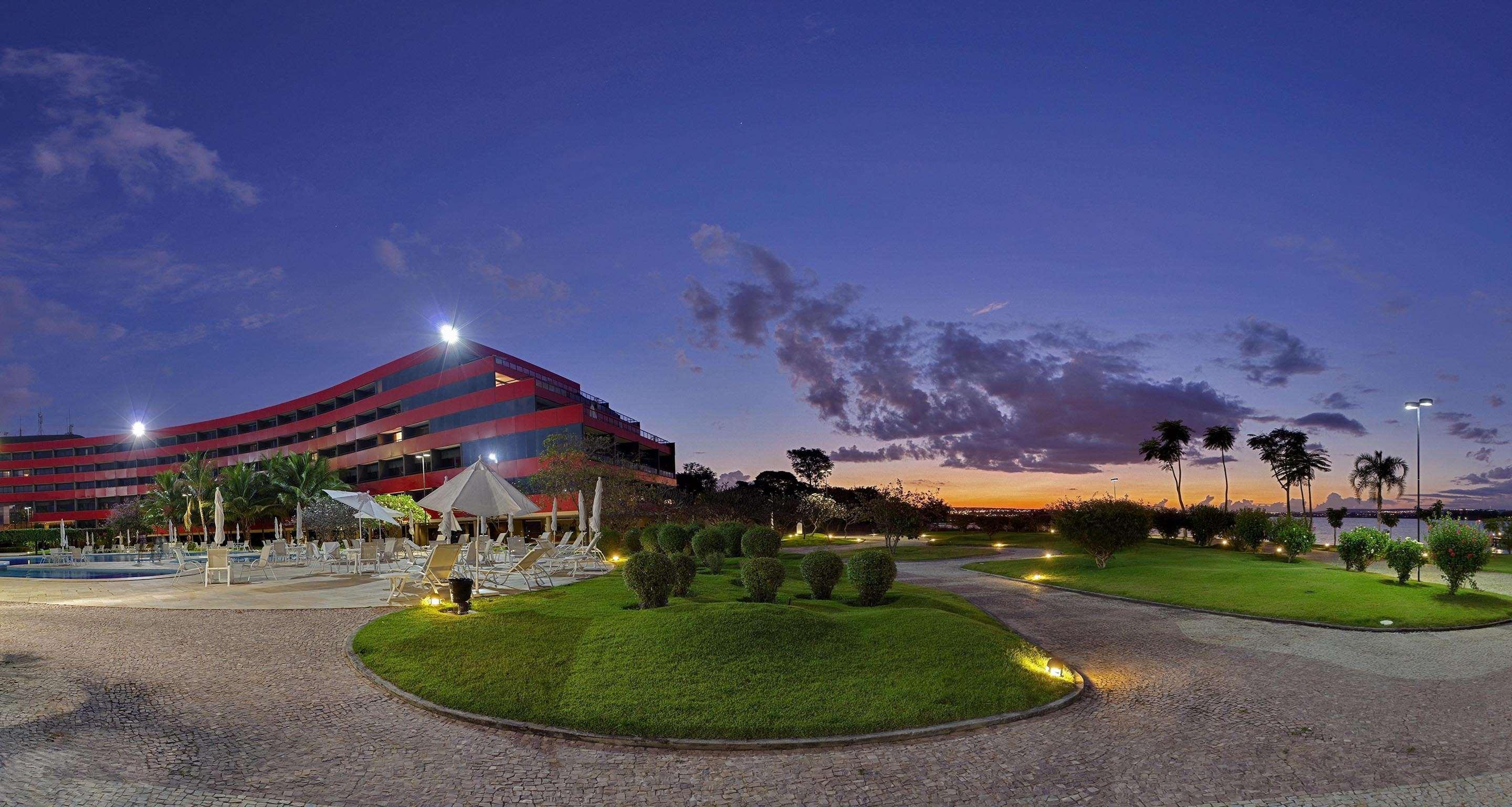 Royal Tulip Brasilia Alvorada Hotel Exterior photo The photo depicts a modern building, possibly a hotel or resort, with a striking red exterior. It is set in a landscaped area that features well-maintained greenery and ornamental plants. In the foreground, there are lounge areas with umbrellas, sugg