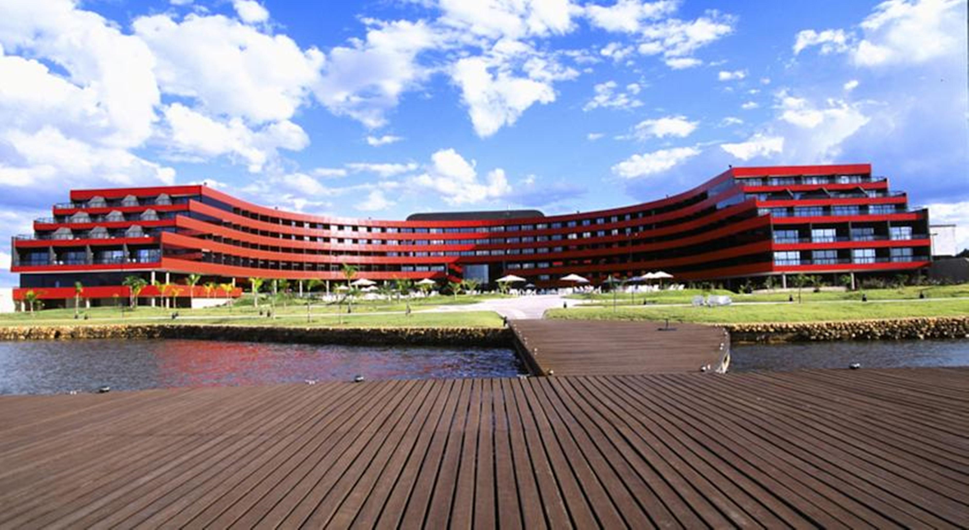 Royal Tulip Brasilia Alvorada Hotel Exterior photo The image shows a modern, curved building with a striking red facade, situated near a body of water. The architecture features multiple levels with large windows that create a sleek and contemporary appearance. In front of the building, there is a wo