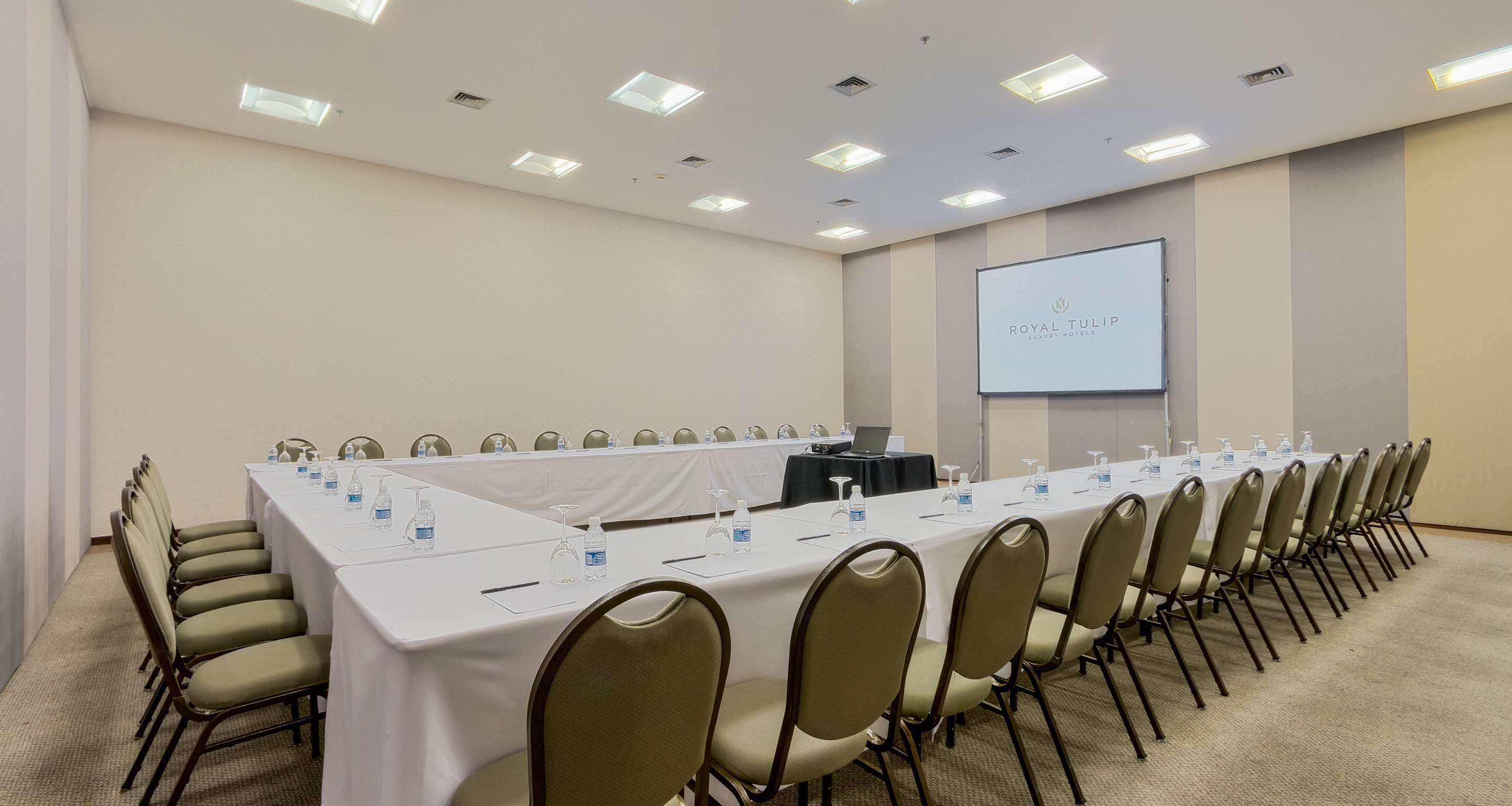 Royal Tulip Brasilia Alvorada Hotel Exterior photo The photo shows a conference room set up for a meeting. There are several tables arranged in a U-shape, covered with white tablecloths. Each table has a water bottle and a notepad, indicating preparation for participants. In the center, there is a sm