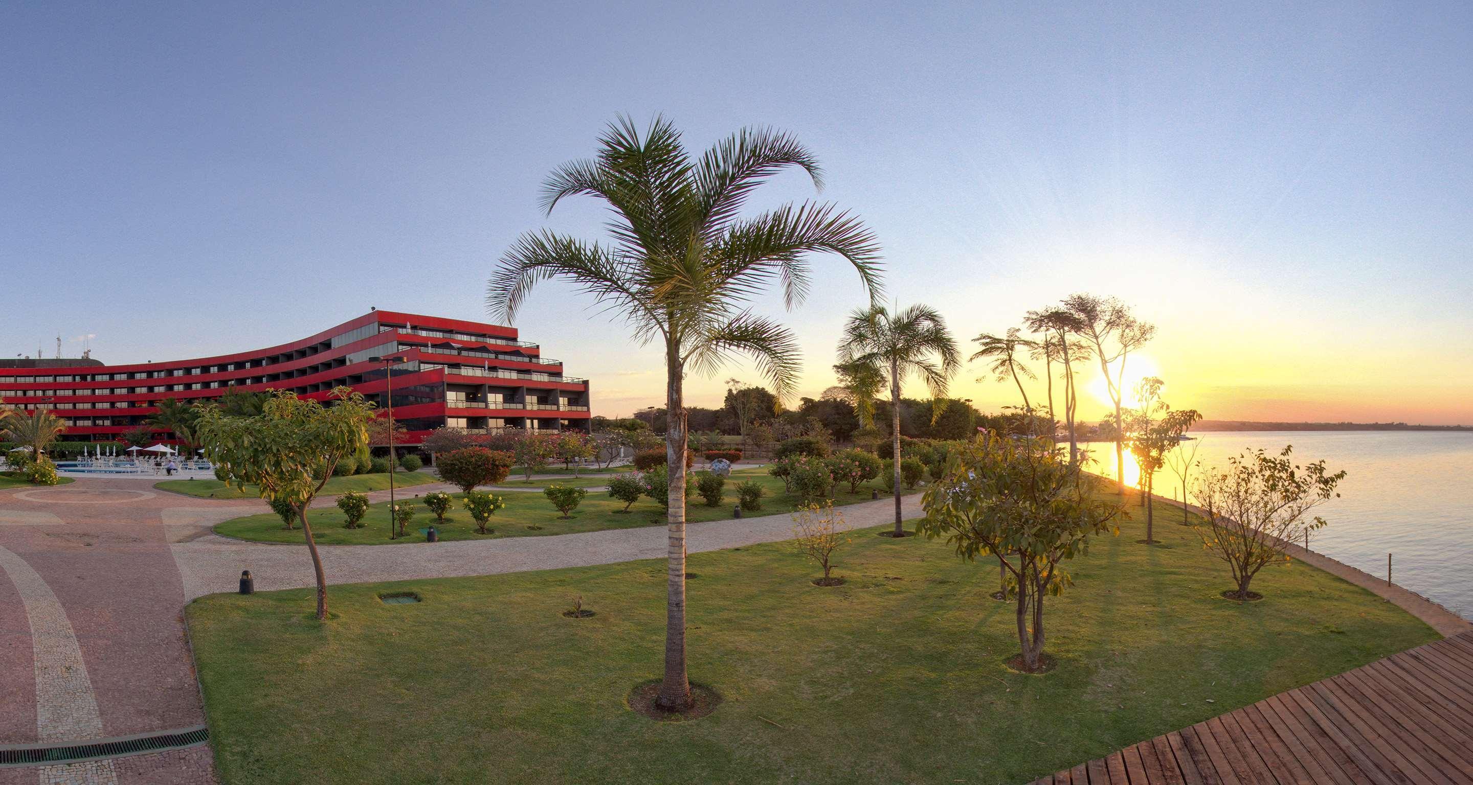 Royal Tulip Brasilia Alvorada Hotel Exterior photo The photo depicts a scenic view featuring a red multi-story building alongside a serene waterfront. In the foreground, there are several palm trees and lush green lawns, creating a tropical atmosphere. The sun is setting in the background, casting wa