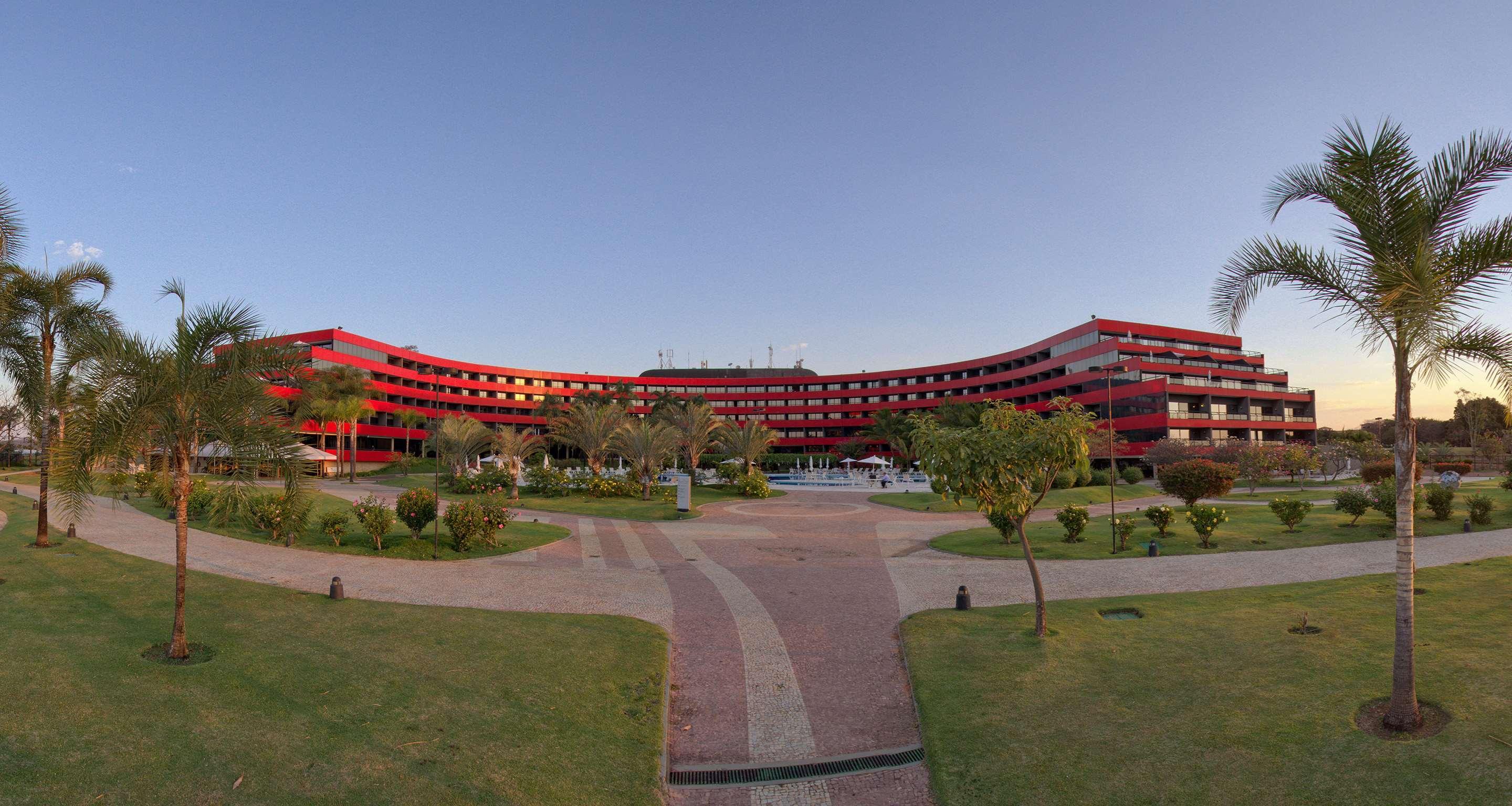 Royal Tulip Brasilia Alvorada Hotel Exterior photo The photo shows a large, curved building with a vibrant red exterior. It appears to be a hotel or resort, surrounded by well-maintained gardens and green spaces. The landscape includes manicured lawns, small trees, and pathways, suggesting a pleasant