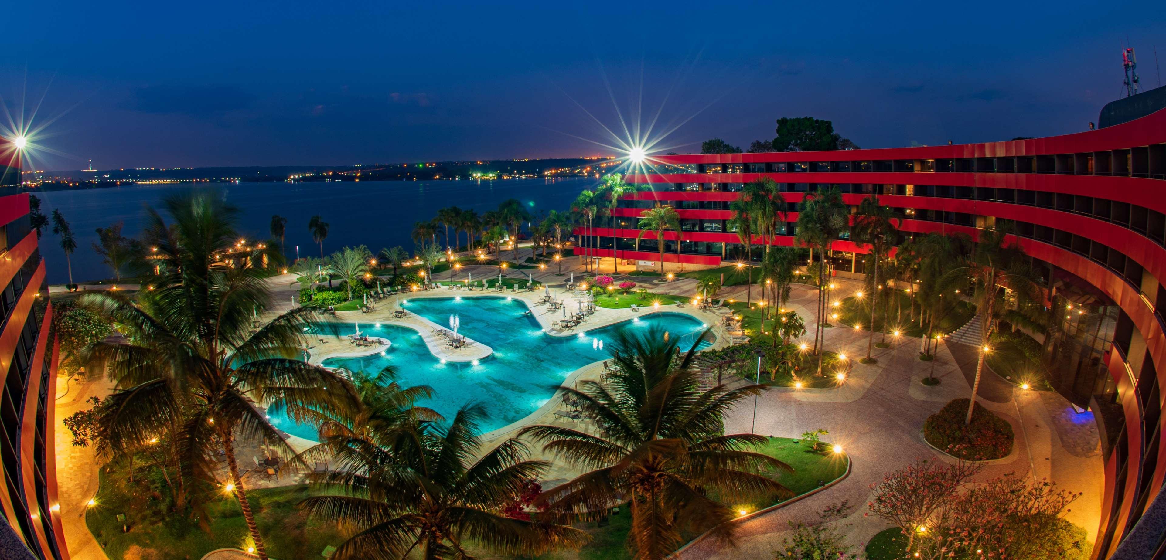 Royal Tulip Brasilia Alvorada Hotel Exterior photo The photo depicts a luxurious resort at night, featuring a large, illuminated swimming pool surrounded by palm trees. The pool has unique shapes and is beautifully lit, creating a tranquil atmosphere. In the background, there is a striking red buildi