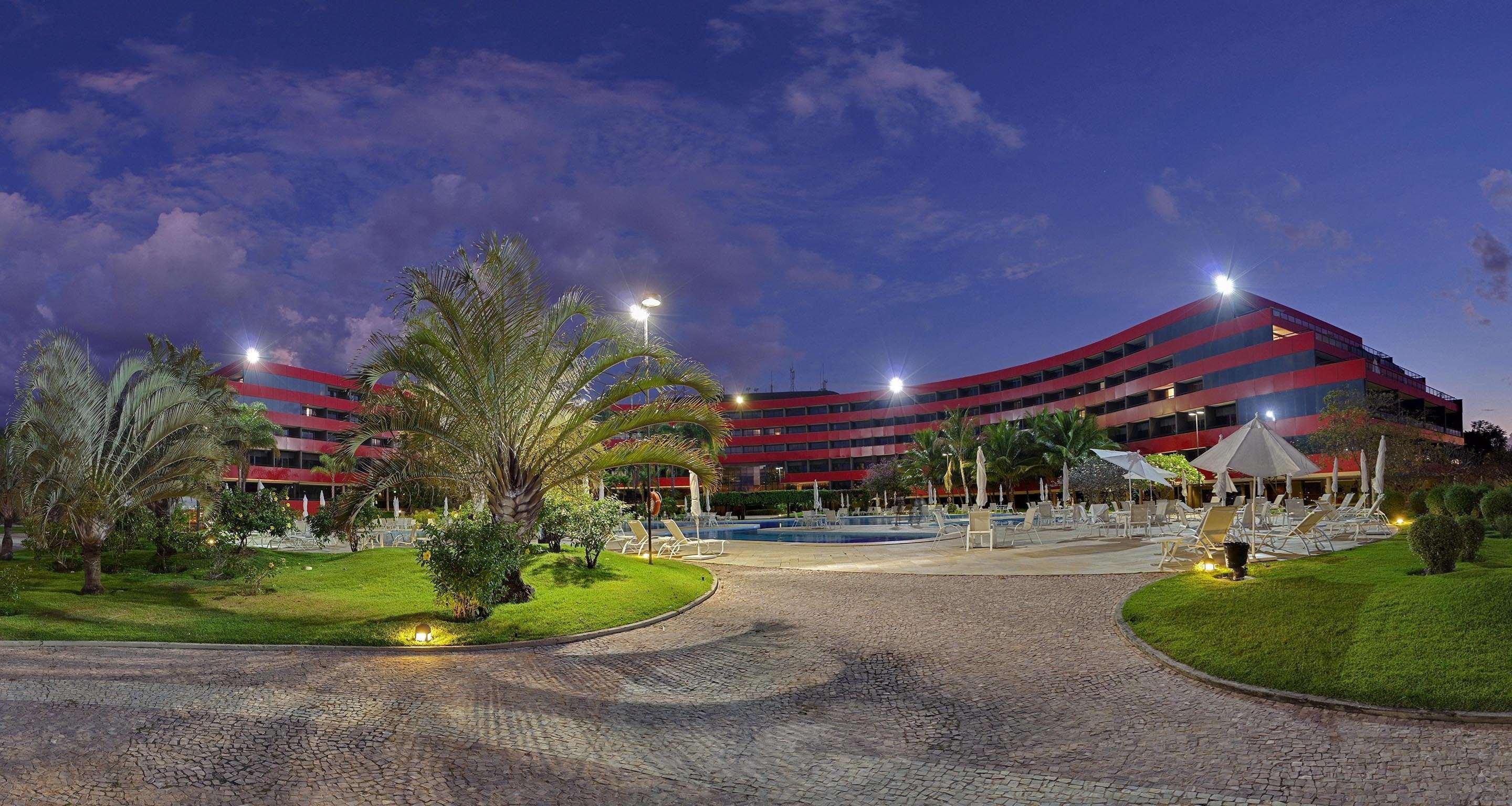 Royal Tulip Brasilia Alvorada Hotel Exterior photo The photo shows a panoramic view of a hotel or resort complex, characterized by its modern architectural design with smooth, curved lines and a vibrant red color. It’s set in a landscaped area featuring palm trees and green grass. The surroundings in