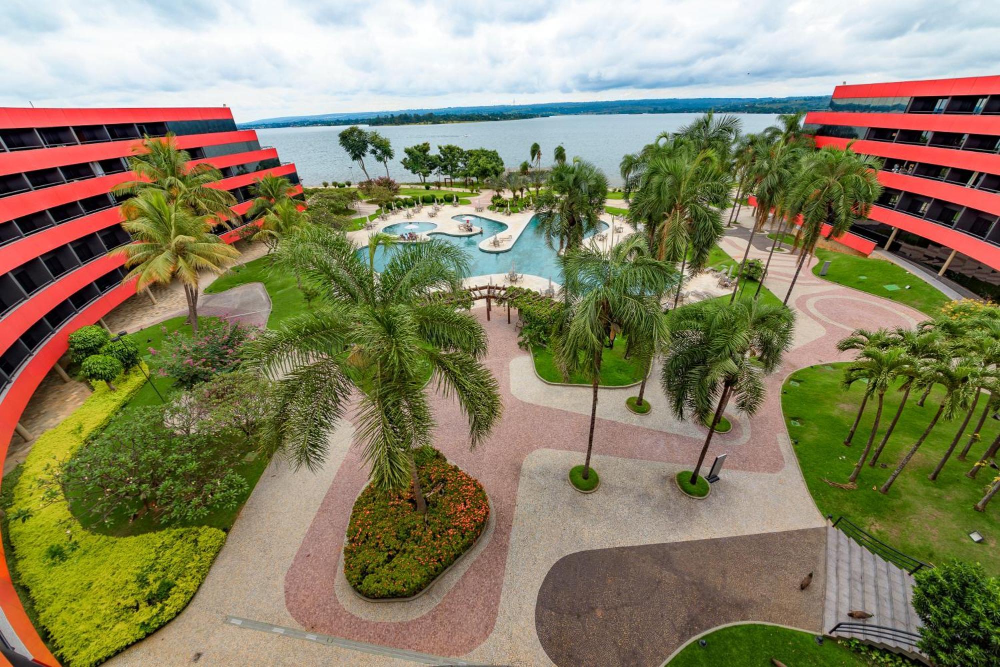 Royal Tulip Brasilia Alvorada Hotel Exterior photo The photo shows a scenic view of a courtyard area in a residential complex. There are multiple red buildings surrounding a pool area, which is surrounded by palm trees and landscaped gardens. The pool features a curvy design, and there are pathways m